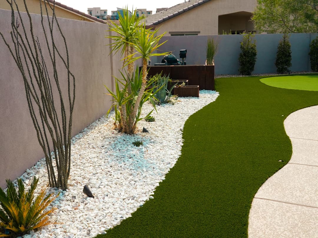 Desert landscape featuring artificial grass, cacti, and decorative white stones in a backyard.