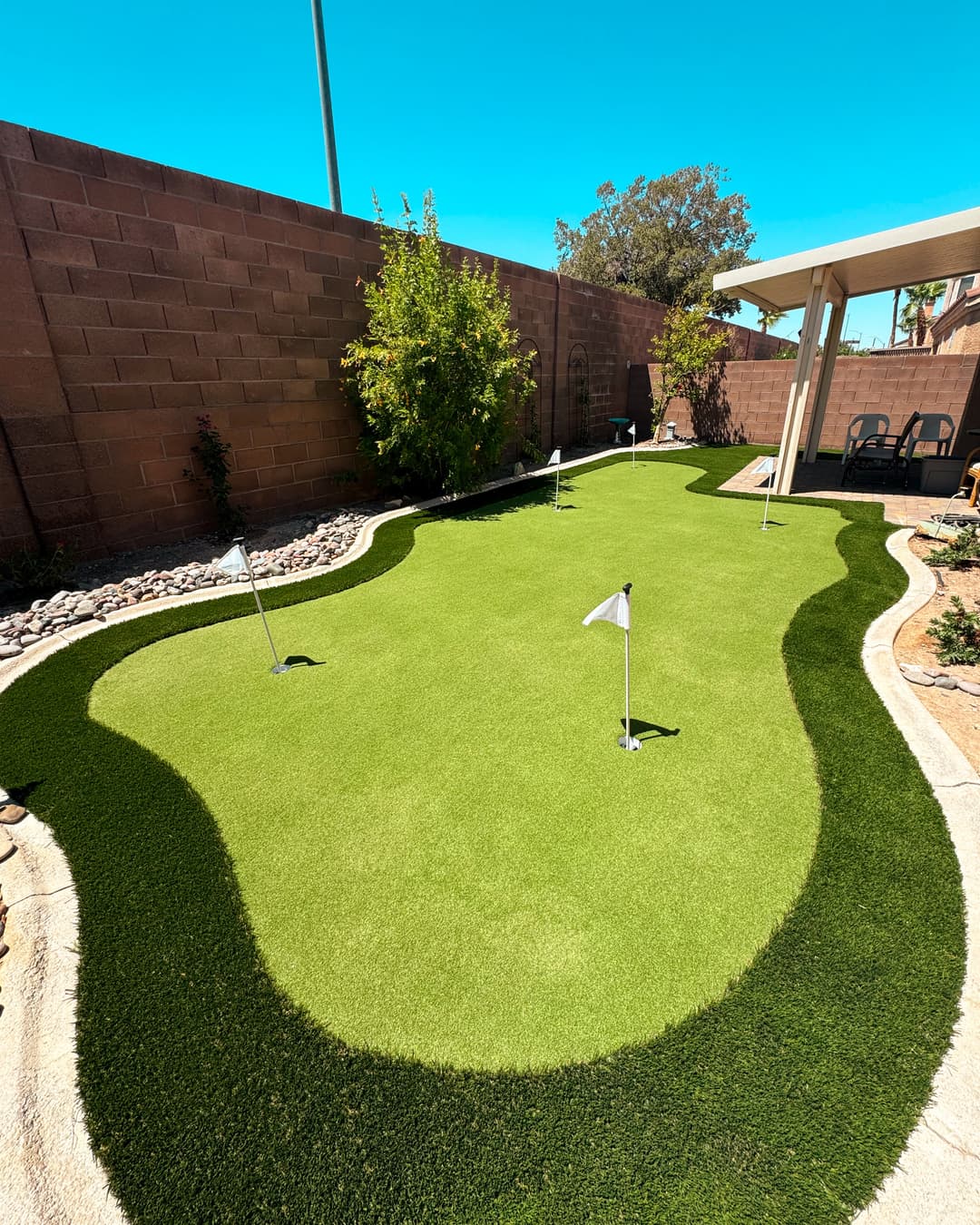 Backyard putting green with flags, surrounded by landscaping and blue sky. Ideal for golf practice.