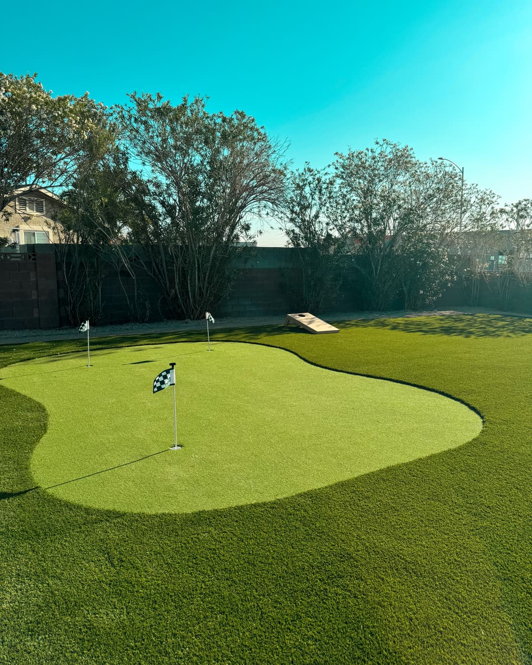 Backyard artificial putting green with flags and a cornhole setup under a clear blue sky.