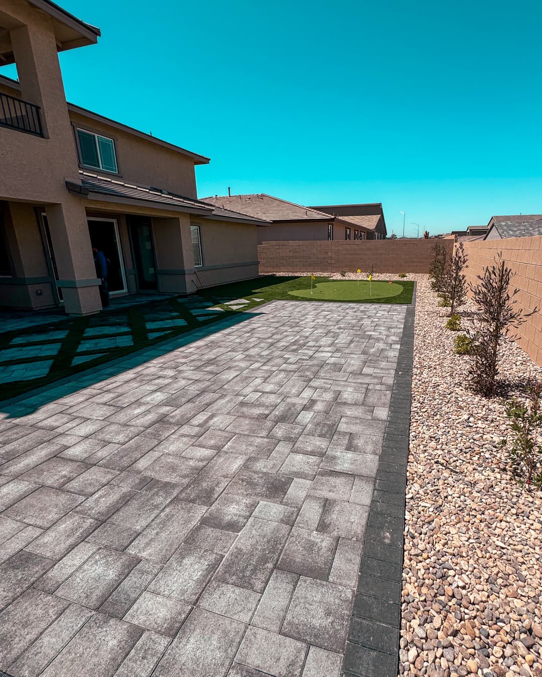 Modern backyard with paved patio, green lawn, and landscaped gravel areas under clear blue sky.