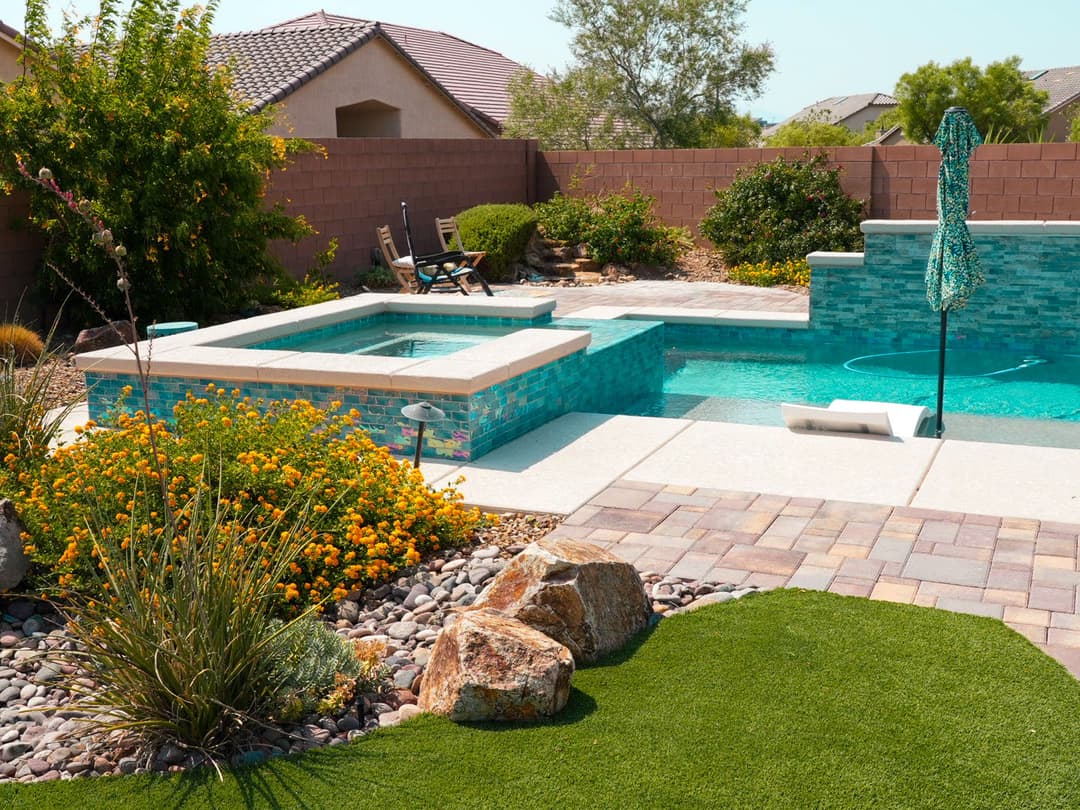 Modern backyard oasis with a turquoise pool, spa, vibrant flowers, and stone landscaping.