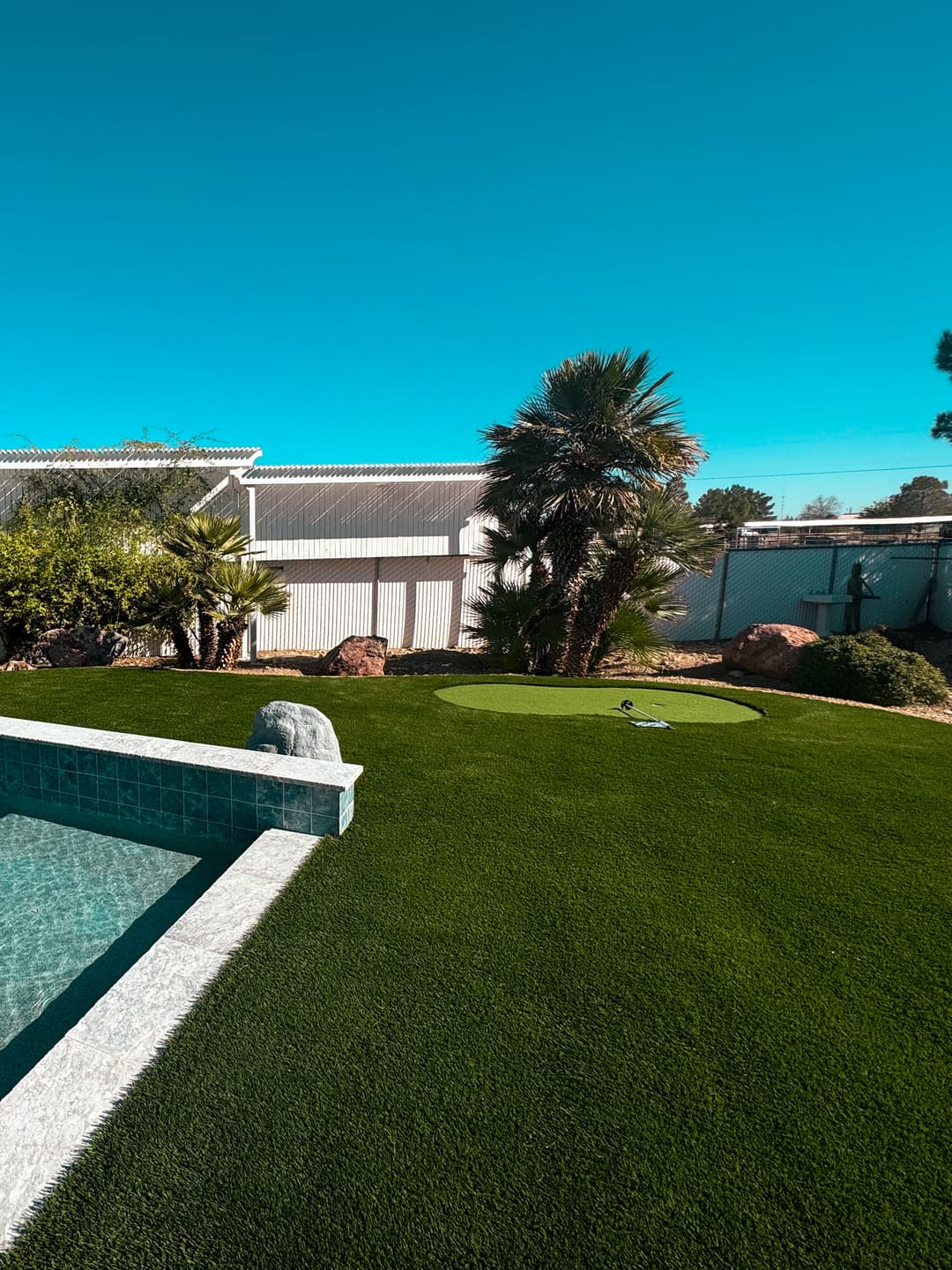 Lush green lawn with a golf putting green beside a swimming pool and palm trees.