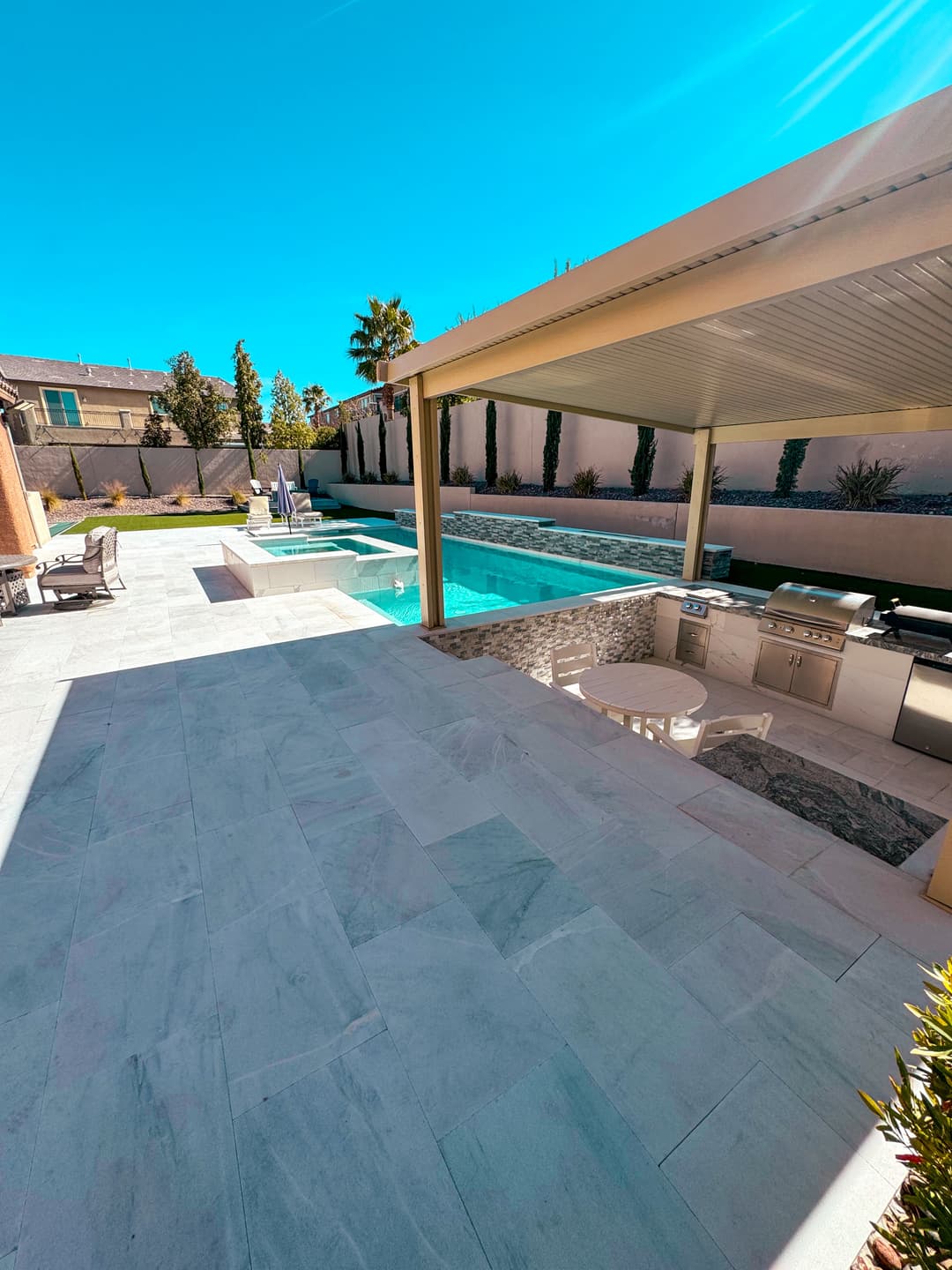 Modern outdoor pool area with cabana, BBQ grill, and lounge chairs under bright blue sky.