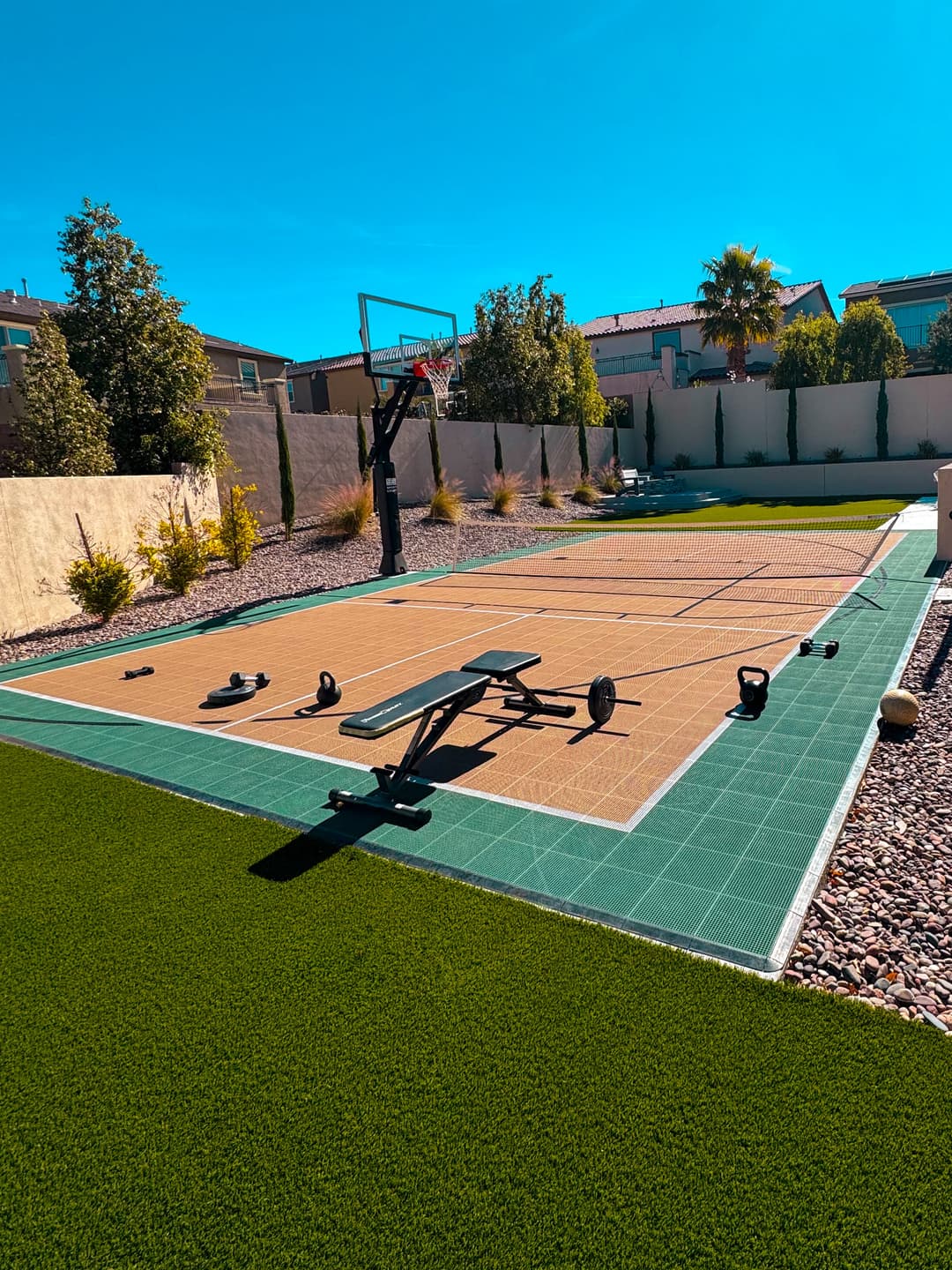 Outdoor basketball court with fitness equipment, surrounded by greenery and bright blue sky.