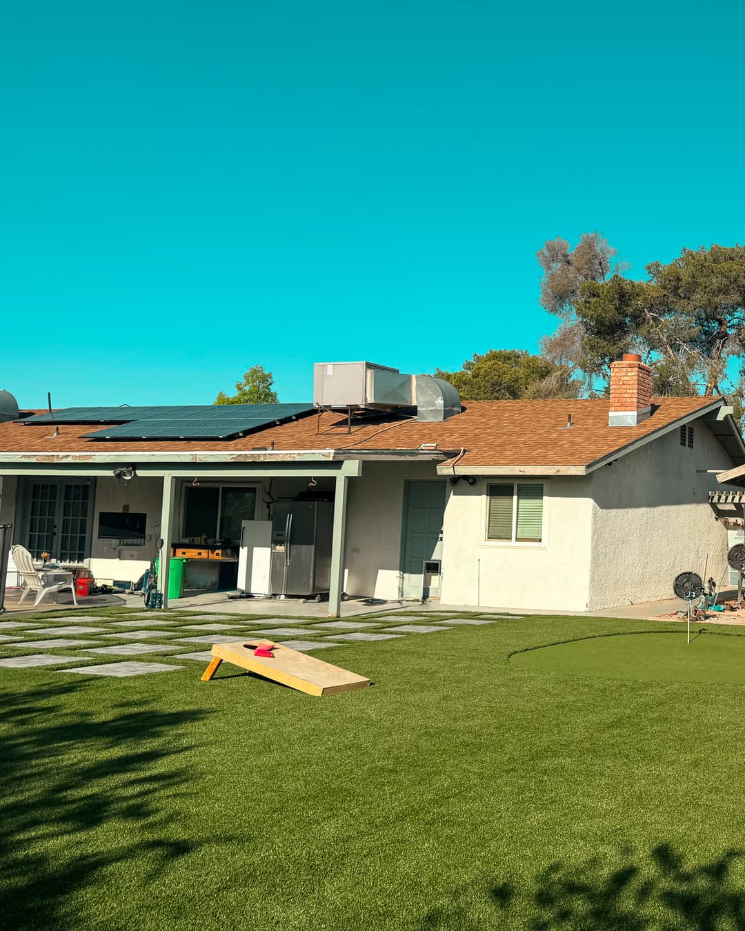 Sunny backyard with artificial turf, patio, and cornhole game setup beside a house.