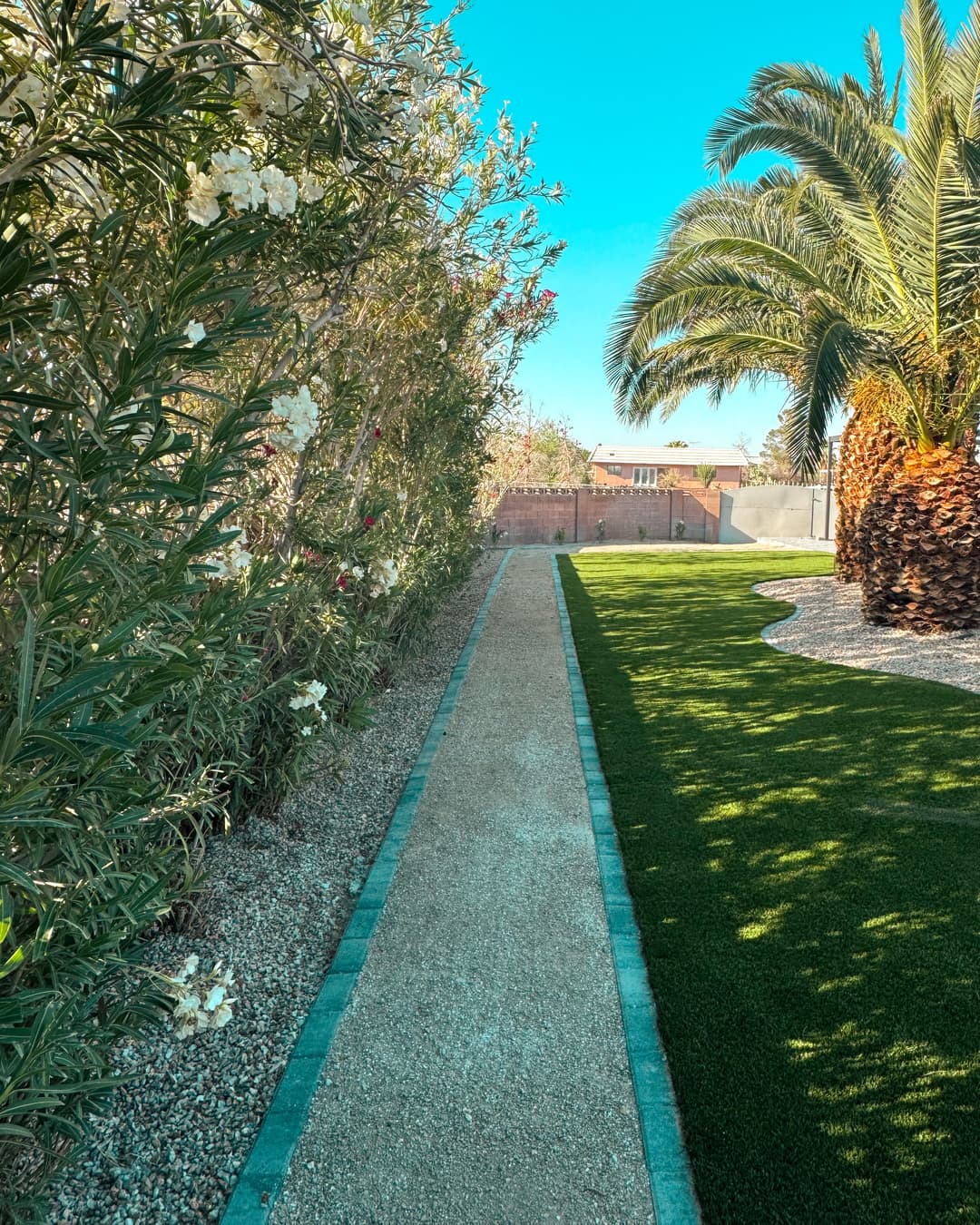 Lush garden pathway lined with flowers, palm trees, and green artificial grass under a blue sky.