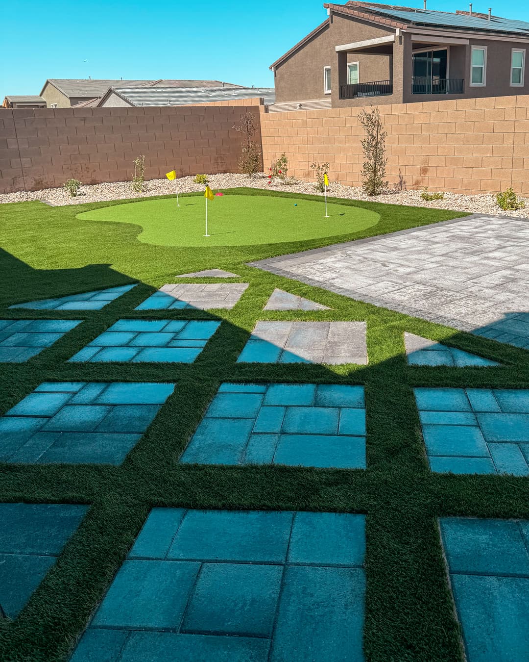 Backyard with manicured synthetic grass, putting green, and paved pathways. Clear blue sky.