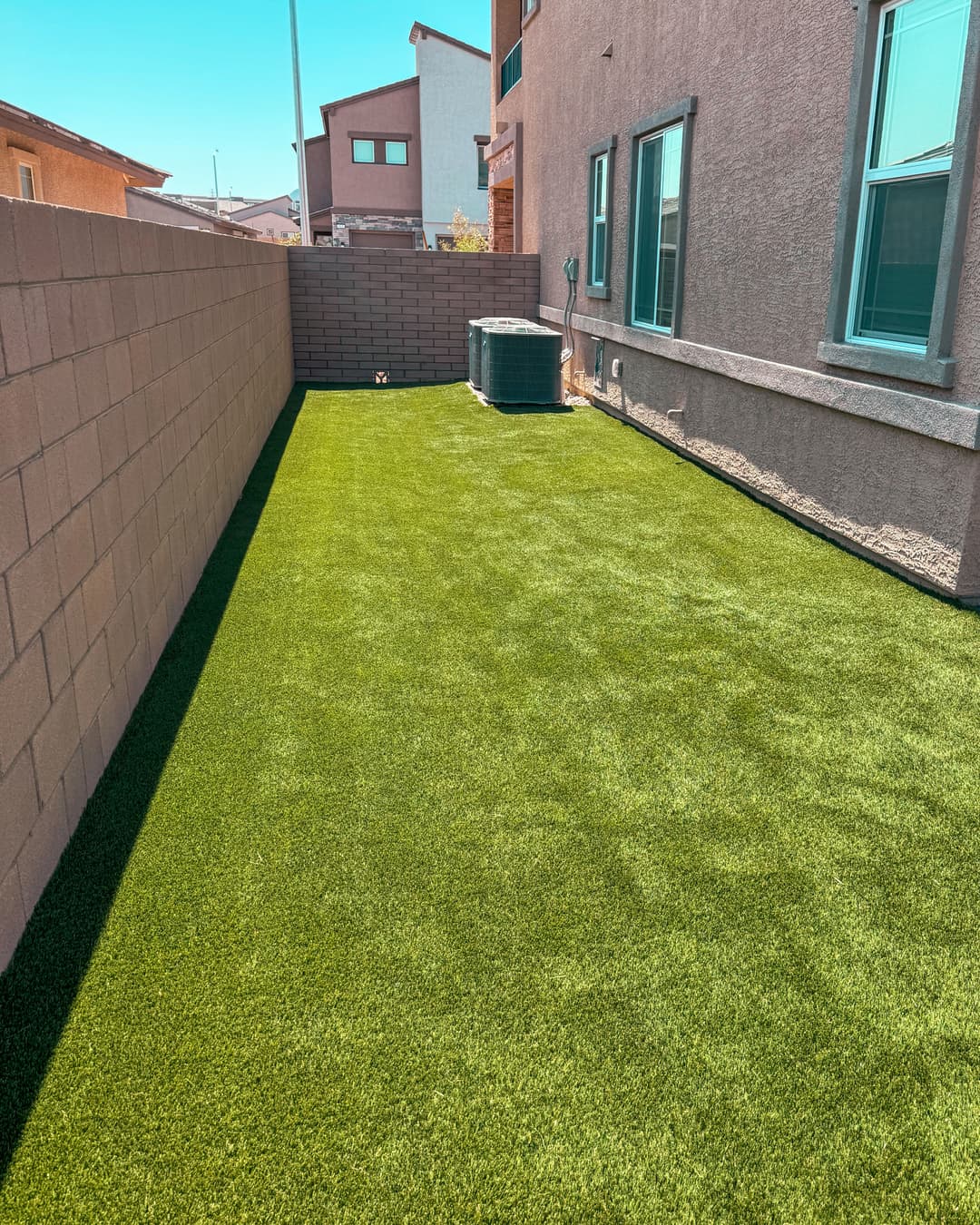 Lush green artificial grass yard between two houses, sunny day, clear blue sky.