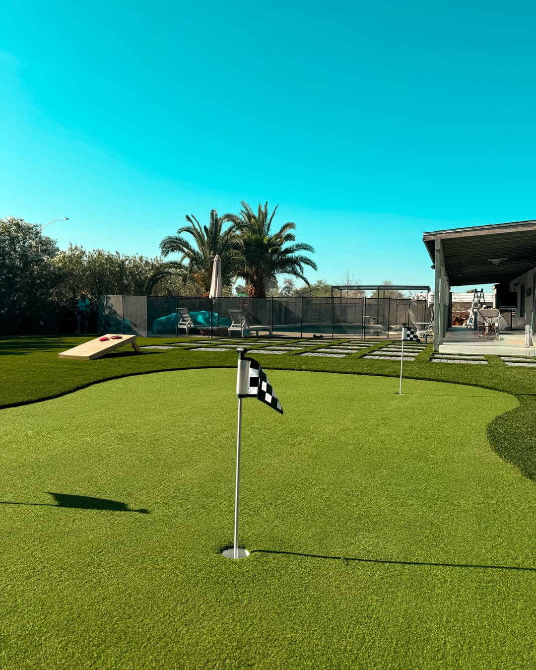 Green backyard putting green with golf flags, palm trees, and lounging area under blue sky.