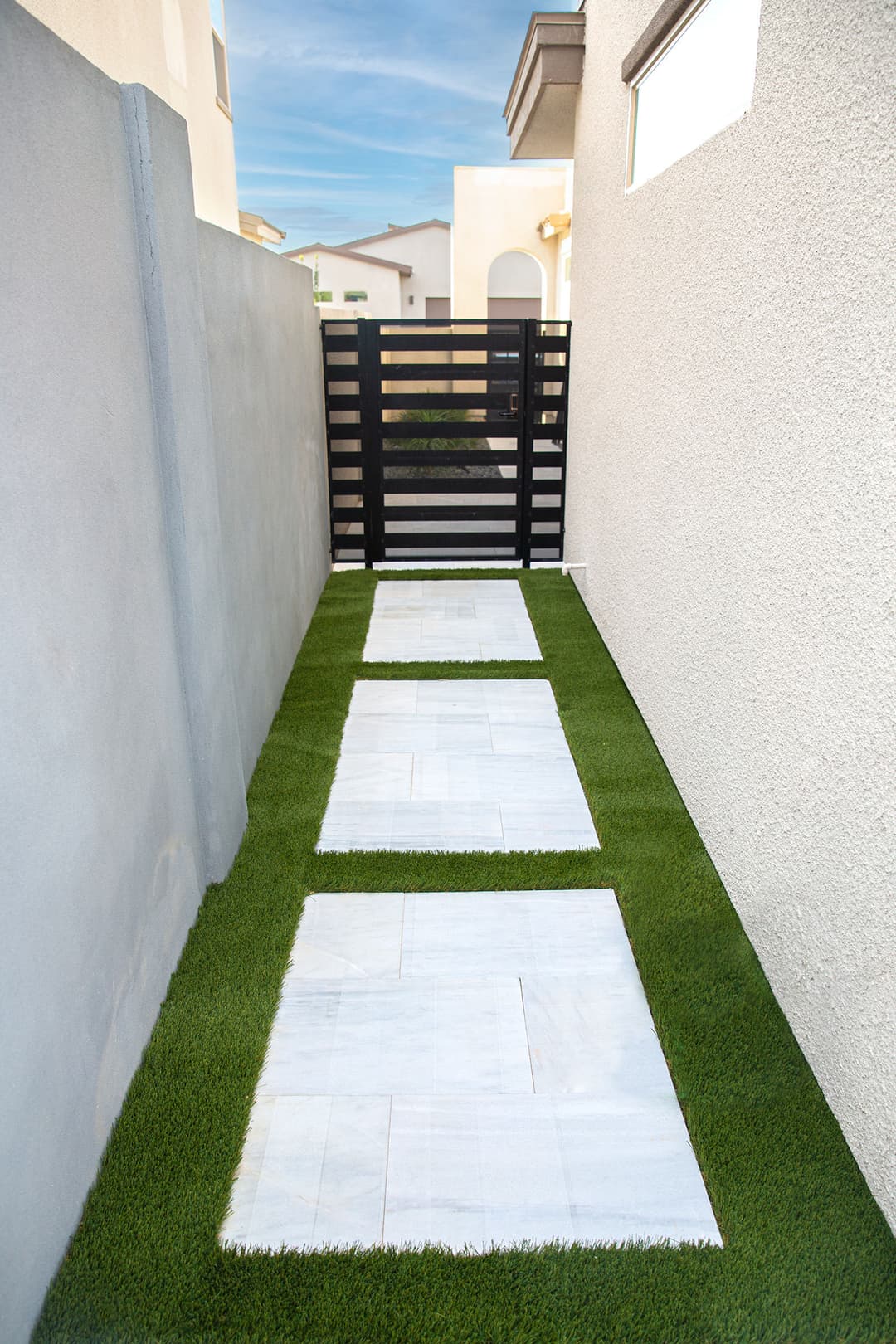 Modern outdoor pathway with white stone slabs and artificial grass, leading to a black wooden gate.